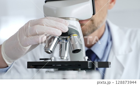 Close up of male scientific researcher wearing white protective gloves carefully calibrating laboratory microscope, performing precision medical investigation. Medicine concept 128873944