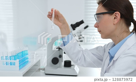 Female researcher examining clear liquid in test tube, sitting near microscopes and lab equipment within sterile scientific workspace. Medicine, healthcare and science concept 128873972