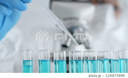 Scientific glassware containing blue solution, researcher carefully dispensing liquid via pipette near microscope within sterile laboratory setting, close up. Medicine, healthcare and science concept Scientific glassware containing blue solution, researcher carefully dispensing liquid via pipette near microscope within sterile laboratory setting, close up. Medicine, healthcare and science concept 128873990