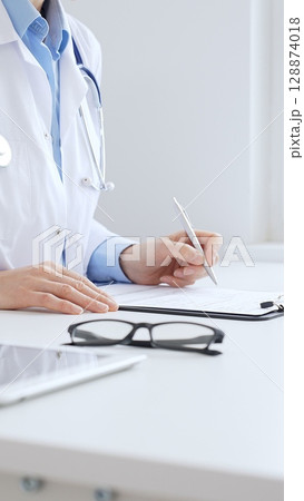 Unknown female doctor completing healthcare paperwork, clipboard positioned near laptop and stethoscope in well lit clinical workspace by window. Medicine and health care Unknown female doctor completing healthcare paperwork, clipboard positioned near laptop and stethoscope in well lit clinical workspace by window. Medicine and health care 128874018
