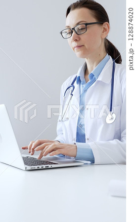Female doctor is typing on a laptop in a bright medical office, showcasing healthcare technology and medical documentation. Medicine and health care concept 128874020