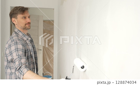 Man professional painter wearing checkered shirt, is rolling white paint on apartment wall with paint roller, transforming interior space with steady strokes during comprehensive home renovation Man professional painter wearing checkered shirt, is rolling white paint on apartment wall with paint roller, transforming interior space with steady strokes during comprehensive home renovation 128874034