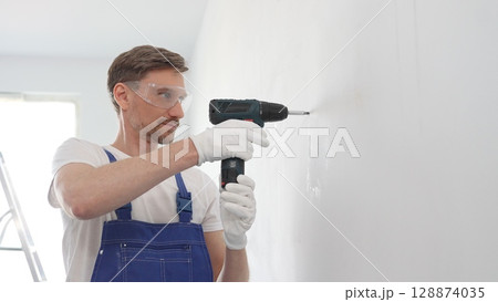 Male construction worker in a blue jumpsuit, safety goggles, and work gloves is screwing a screw into the wall using a cordless screwdriver, carrying out a careful home renovation with professional 128874035