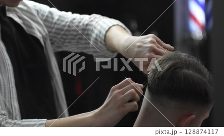 Skilled barber trimming customer's hair using professional scissors and comb, working in contemporary barbershop with vintage pole visible near workspace Skilled barber trimming customer's hair using professional scissors and comb, working in contemporary barbershop with vintage pole visible near workspace 128874117