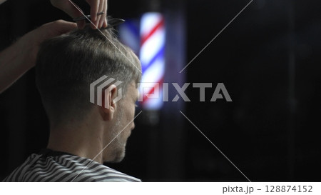 Professional barber cutting hair of male client in barbershop, using scissors and comb, with barber pole in background, creating stylish and modern haircut 128874152