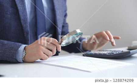 Businessman financial analyst is analyzing financial documents with a calculator and magnifying glass, ensuring accuracy and compliance in the office background. Audit and taxes in business Businessman financial analyst is analyzing financial documents with a calculator and magnifying glass, ensuring accuracy and compliance in the office background. Audit and taxes in business 128874209