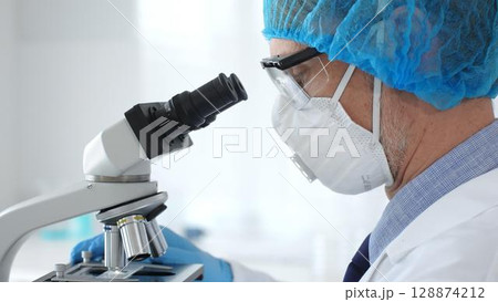 Male scientist wearing protective gear examining microscope slide, analyzing research sample in sterile laboratory environment, close up. Medicine and science concept Male scientist wearing protective gear examining microscope slide, analyzing research sample in sterile laboratory environment, close up. Medicine and science concept 128874212