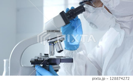 Medical virologist in a hazmat suit, mask, and gloves, diligently working at a microscope in a laboratory researching viruses. Healthcare and science concept 128874272
