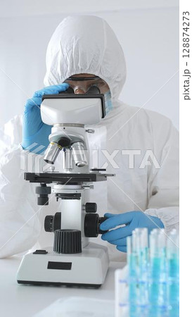 A medical virologist in protective gear examines viral samples under a microscope while conducting research in a laboratory setting 128874273