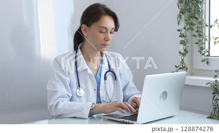 Focused female doctor wearing lab coat and stethoscope, sitting at desk and working on her laptop in bright medical office with window and decorative plants. Medicine and health care 128874276