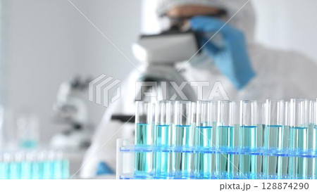 Male medical scientist in protective suit using microscope while analyzing samples, conducting research in laboratory. Close-up of test tubes with blue liquid Male medical scientist in protective suit using microscope while analyzing samples, conducting research in laboratory. Close-up of test tubes with blue liquid 128874290