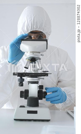 A medical virologist in protective gear examines viral samples under a microscope while conducting research in a laboratory setting 128874302