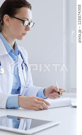 Adult doctor woman completing healthcare paperwork, clipboard positioned near laptop and stethoscope in well lit clinical workspace by window. Medicine and health care Adult doctor woman completing healthcare paperwork, clipboard positioned near laptop and stethoscope in well lit clinical workspace by window. Medicine and health care 128874308