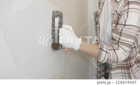 Unknown female construction worker wearing protective gloves applying plaster, smoothing wall surface during home renovation work Unknown female construction worker wearing protective gloves applying plaster, smoothing wall surface during home renovation work 128874407