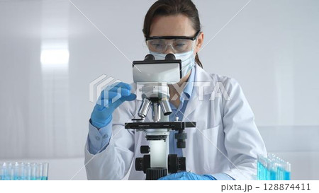 A female scientist in a lab coat and blue gloves examines a microscope slide in a sterile laboratory, conducting research using blue samples from a test tube 128874411