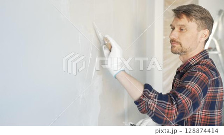 Male construction worker spreading plaster on white wall, wearing protective gloves during home renovation project with putty knife 128874414