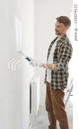 Professional house painter wearing gloves painting a white wall with a paint roller, working on a home renovation project, creating a fresh and clean look Professional house painter wearing gloves painting a white wall with a paint roller, working on a home renovation project, creating a fresh and clean look 128874415