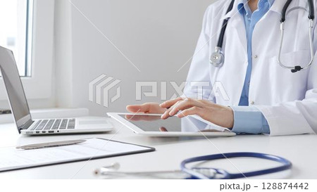 Female doctor wearing white medical coat and stethoscope using digital tablet with laptop and medical records on desk in medical office, showcasing modern healthcare technology. Medicine and helthcare 128874442