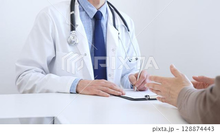 Adult aged doctor man taking notes while listening to a female patient explaining her symptoms during a medical consultation in the clinic office. Medicine and health care concept Adult aged doctor man taking notes while listening to a female patient explaining her symptoms during a medical consultation in the clinic office. Medicine and health care concept 128874443