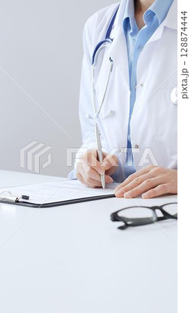 Unknown female doctor completing healthcare paperwork, clipboard positioned near laptop and stethoscope in well lit clinical workspace by window. Medicine and health care 128874444