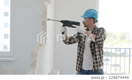 Man construction worker demolishing a wall with a hammer drill in protective gear, creating construction debris on the surrounding floor surface. Renovation concept 128874449