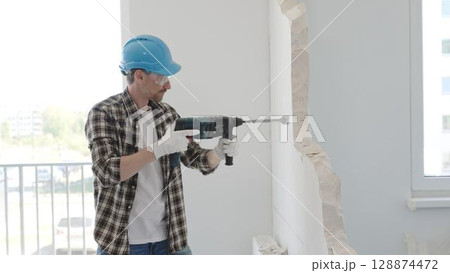 Man construction worker demolishing a wall with a hammer drill in protective gear, creating construction debris on the surrounding floor surface. Renovation concept 128874472