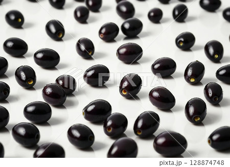 Various black olives are scattered playfully on a clean white background, their glossy surfaces catching the light, highlighting their rich, deep hues and unique shapes Generative AI 128874748