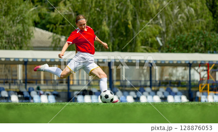 Woman football player in red jersey kicking soccer ball on green field 128876053