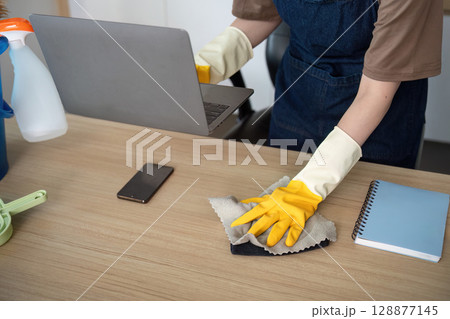 Home Cleaning. Young woman with gloves cleaning her desk while using a laptop. Home Cleaning. Young woman with gloves cleaning her desk while using a laptop. 128877145