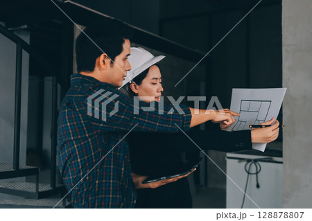 Construction manager and engineer dressed in orange work vests and hard helmets explore construction documentation on the building site near the steel frames 128878807