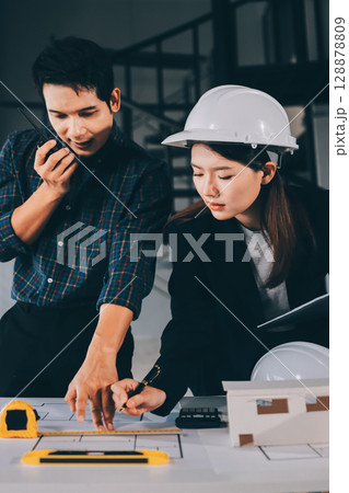 Construction manager and engineer dressed in orange work vests and hard helmets explore construction documentation on the building site near the steel frames 128878809