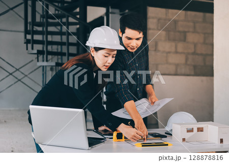 Construction manager and engineer dressed in orange work vests and hard helmets explore construction documentation on the building site near the steel frames 128878816