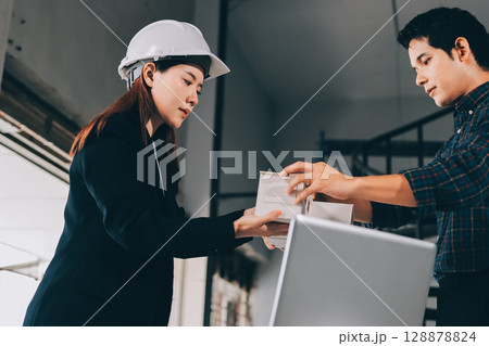 Construction manager and engineer dressed in orange work vests and hard helmets explore construction documentation on the building site near the steel frames 128878824