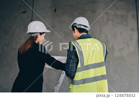 Construction manager and engineer dressed in orange work vests and hard helmets explore construction documentation on the building site near the steel frames Construction manager and engineer dressed in orange work vests and hard helmets explore construction documentation on the building site near the steel frames 128878832