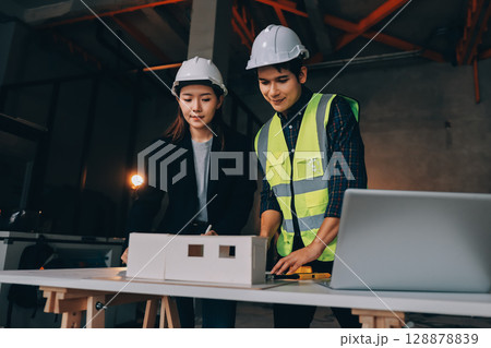 Construction manager and engineer dressed in orange work vests and hard helmets explore construction documentation on the building site near the steel frames Construction manager and engineer dressed in orange work vests and hard helmets explore construction documentation on the building site near the steel frames 128878839