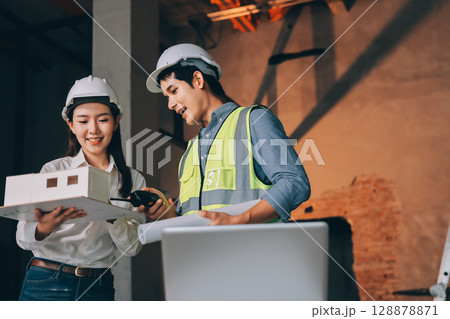 Construction manager and engineer dressed in orange work vests and hard helmets explore construction documentation on the building site near the steel frames Construction manager and engineer dressed in orange work vests and hard helmets explore construction documentation on the building site near the steel frames 128878871