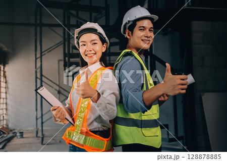 Construction manager and engineer dressed in orange work vests and hard helmets explore construction documentation on the building site near the steel frames 128878885
