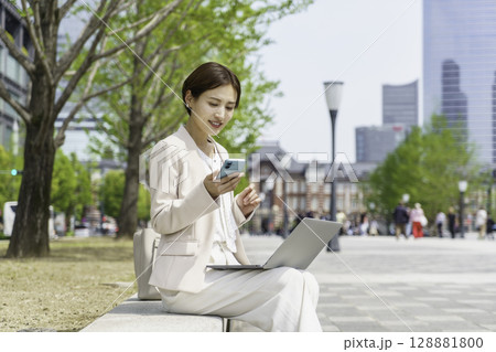 ベンチに座ってパソコンを見ながらスマホを操作する若いビジネスウーマン ベンチに座ってパソコンを見ながらスマホを操作する若いビジネスウーマン 128881800