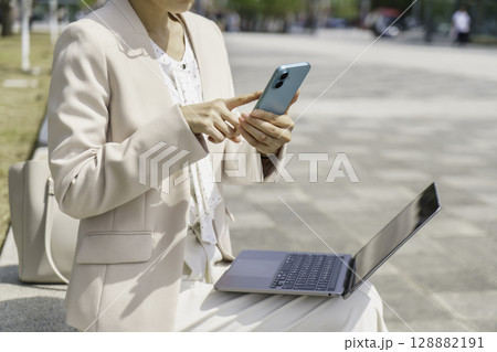 ベンチに座ってノートパソコンを見ながらスマホを操作する若いビジネスウーマンの手元のアップ 128882191