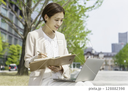 ベンチに座ってノートパソコンを見ながら手帳に書き込みをする若いビジネスウーマン 128882398