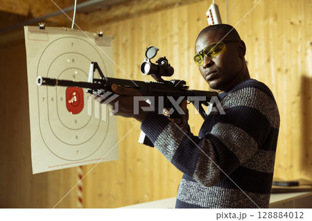 Portrait of man holding rifle enjoying fun indoor shooting experience in firing range. Person wearing protective equipment practicing with semiautomatic weapon in safe, welcoming environment 128884012