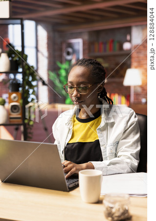 Student at home browsing internet on notebook, doing homework, researching information online. African american girl in apartment reading articles on laptop for school tasks 128884044