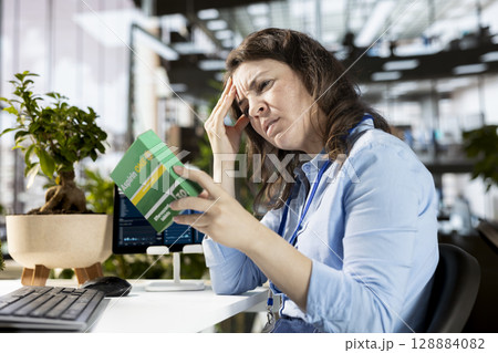 Businesswoman with a headache preparing to take a painkiller medicine in order to solve her daily organizational tasks, reading the recommended dosage on the box label. Aspirin pills. 128884082
