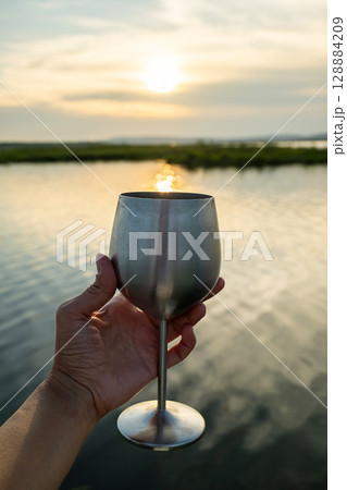 Woman hand holding elegant metal wine glass against scenic lake sunset. 128884209
