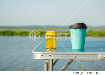 Yellow vitamin container and teal travel mug on blue camping table outdoors by the lake. 128884215