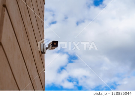 Surveillance Camera on Building Wall Against Cloudy Sky, Security and Control 128886044