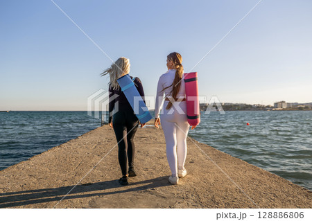 Yoga Women Beach Pier - Two women carrying yoga mats walk along a pier towards the ocean at sunset. 128886806