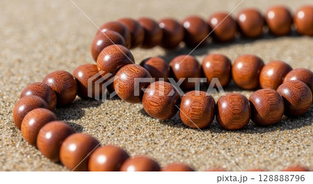Polished wooden prayer beads mala necklace scattered on sandy beach surface with natural sunlight creating warm shadows and reflective highlights 128888796