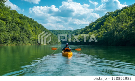Person kayaking down a tranquil green river surrounded by lush forests and verdant hills under a clear blue sky 128891982