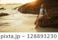 Woman leaning on a rock reef enjoying and relax warm dark golden sunset. Palm tree in beach. Woman in blue swimsuit and sunglasses Woman leaning on a rock reef enjoying and relax warm dark golden sunset. Palm tree in beach. Woman in blue swimsuit and sunglasses 128893230